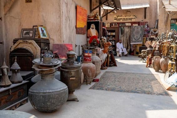 मार्केट दृश्य जिसमें प्राचीन जार, कालीन, चित्र, और पृष्ठभूमि में बैठे एक विक्रेता ग्राहक की प्रतीक्षा कर रहे हैं।