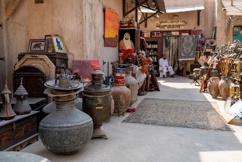 मार्केट दृश्य जिसमें प्राचीन जार, कालीन, चित्र, और पृष्ठभूमि में बैठे एक विक्रेता ग्राहक की प्रतीक्षा कर रहे हैं।