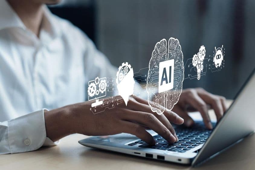 नोटबुक पर प्रक्षेपण जिसमें एक AI मस्तिष्क और अन्य तत्व दिखाए गए हैं। कंप्यूटर पर काम करते हुए एक मानव हाथ।