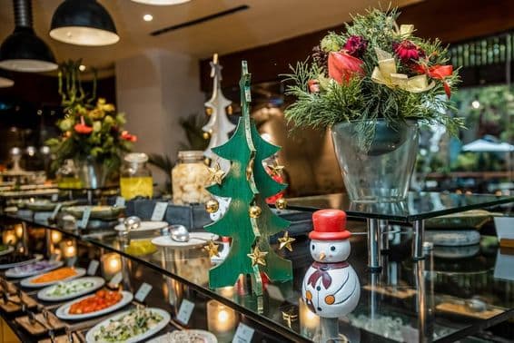 रेस्टोरेंट की सब्जियों की काउंटर पर एक छोटा स्नोमैन, बगल में लकड़ी का क्रिसमस पेड़।