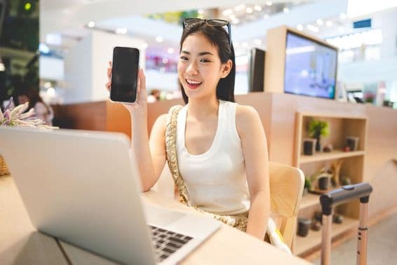 जेनरेशन Z लड़की काम कर रही है, फोन पकड़े हुए, उसके सामने एक नोटबुक और बगल में एक रोलिंग सूटकेस का हैंडल।