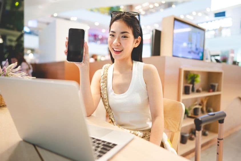 जेनरेशन Z लड़की काम कर रही है, फोन पकड़े हुए, उसके सामने एक नोटबुक और बगल में एक रोलिंग सूटकेस का हैंडल।