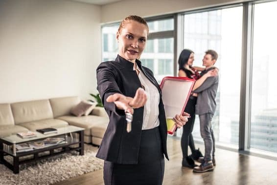 एक महिला रियल एस्टेट ब्रोकर हाथ हिलाती हुए, पीछे एक जोड़ा गले मिलता हुआ।