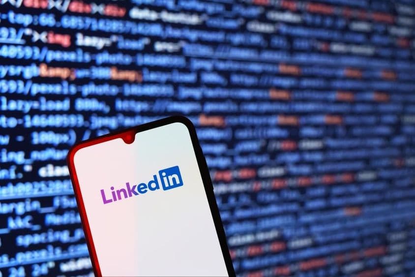 स्मार्टफोन जिसमें LinkedIn लोगो के साथ बैकग्राउंड में कोड स्निपेट दिखाई दे रहा है।