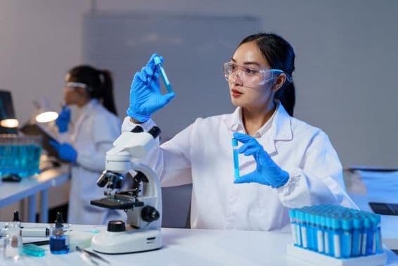 लैब कोट और दस्ताने पहने एशियाई महिला वैज्ञानिक दो टेस्ट ट्यूब की तुलना करते हुए।