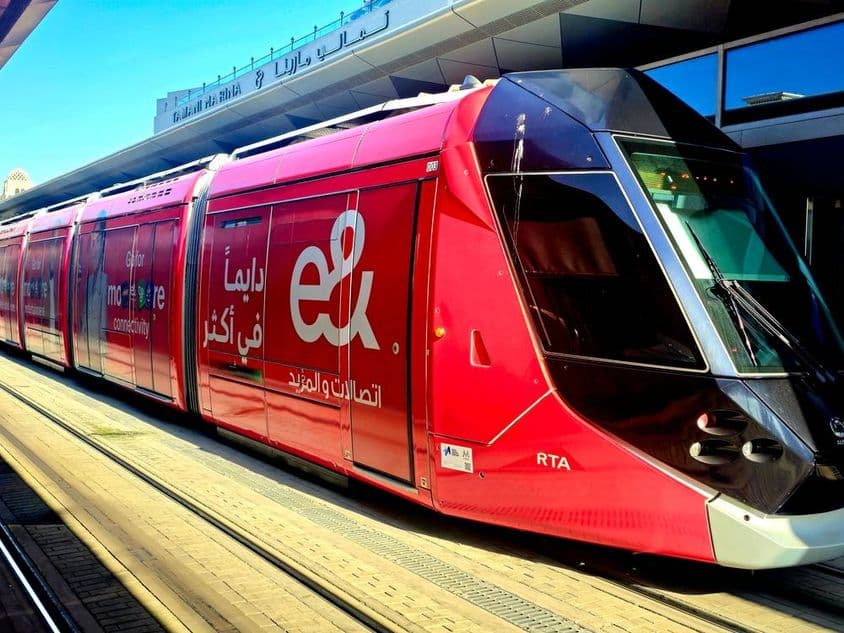 एक प्लेटफॉर्म पर पार्क की गई लाल ट्राम, बड़ी खिड़कियों और सुरुचिपूर्ण डिजाइन के साथ।