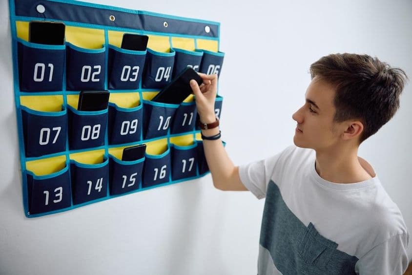 छात्र एक दीवार पर लगे फोन आयोजक में स्मार्टफोन छोड़ता है।