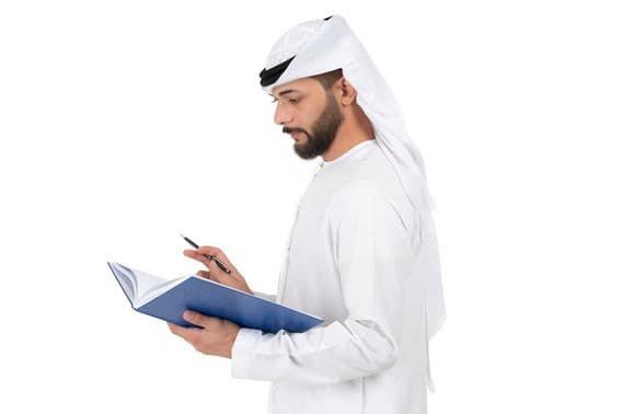 एक युवा अरब शिक्षक खड़े हैं और किताब में लिख रहे हैं।