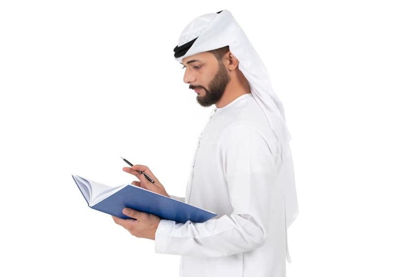 एक युवा अरब शिक्षक खड़े हैं और किताब में लिख रहे हैं।