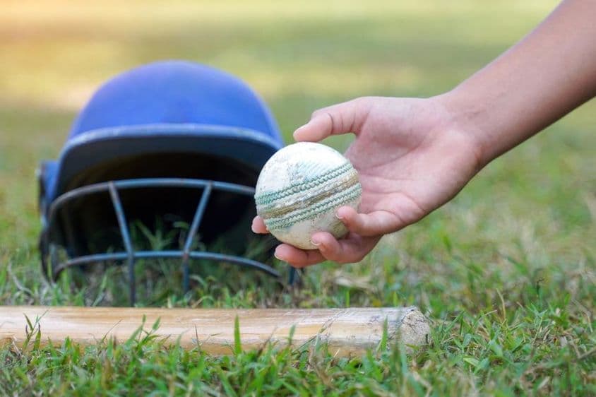 घास पर पड़े अन्य क्रिकेट गियर के साथ एक खिलाड़ी के हाथ का क्रिकेट गेंद उठाते हुए।
