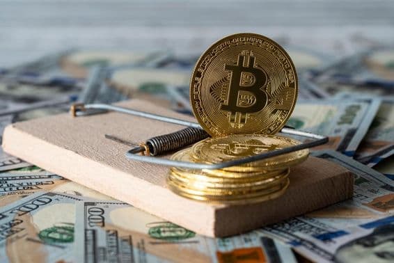 गोल्ड क्रिप्टोकरेंसी सिक्के, बिटकॉइन एक चूहेदानी में, पृष्ठभूमि में अमेरिकी १०० डॉलर के बिल के साथ।